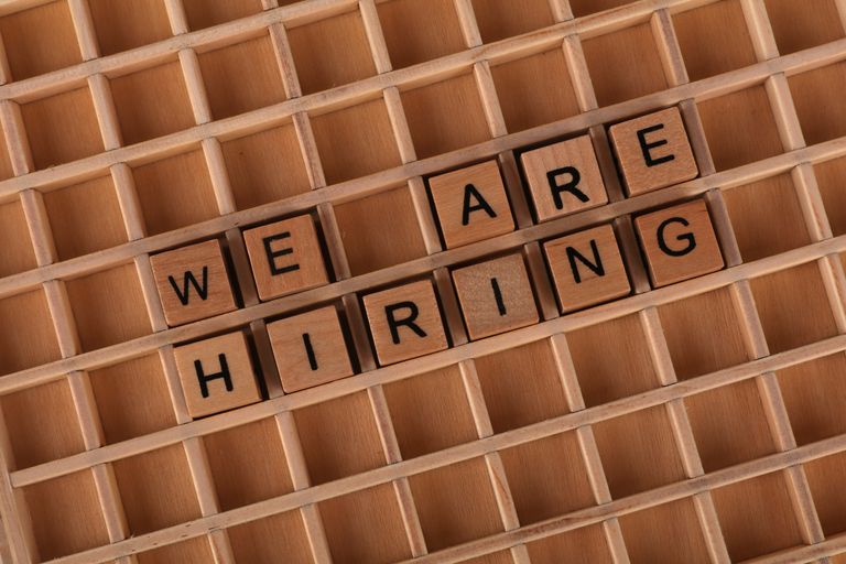 Houten lettertegels die zo zijn gerangschikt dat ze ‘We Are Hiring’ vormen, tegen een houten rasterachtergrond.